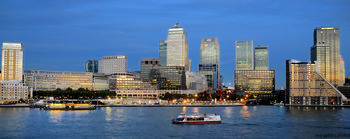 Canary wharf dusk This urban architecture photograph captures the city skyline of Canary Wharf in London, United Kingdom, during a November evening in autumn. The image prominently displays the illuminated modern skyscrapers of Canary Wharf along the north bank of the River Thames. Landmark buildings such as One Canada Square and the Citi building are clearly visible as part of the Canary Wharf financial district. In the foreground, boats are seen moving along the River Thames, adding movement to the urban scene. The evening light provides a blue hue to the sky, enhancing the architectural features of the cityscape and reflecting the vibrant city atmosphere of London.